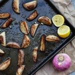 This is a photo of roasted turnips on a dark roasting sheet. There's some lemon in the photo and an uncooked turnip as well in the photo.