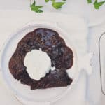 photo of chocolate pudding cake with fresh whipped cream on a white plate on a white cloth. Green vines are in the background and the shot is from above.