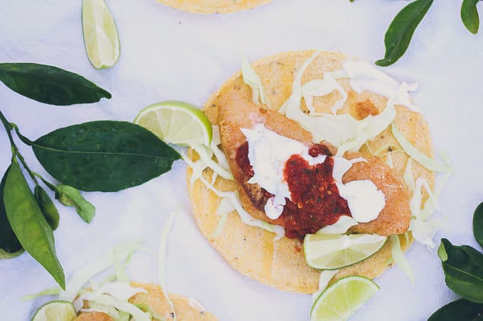 photo of three baja fish tacos on white tablecloth, surrounded by some citrus leaves and limes. Tacos have fist, crema, salsa, and cabbage on top of open tortillas.