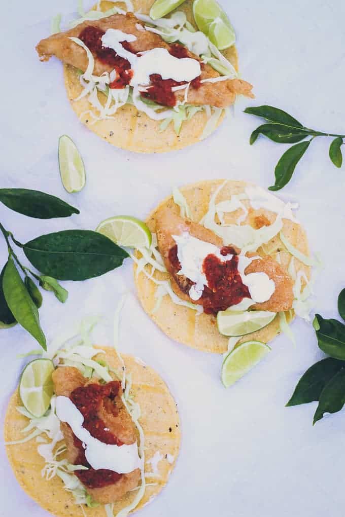 photo of three baja fish tacos on white tablecloth, surrounded by some citrus leaves and limes. Tacos have fist, crema, salsa, and cabbage on top of open tortillas.