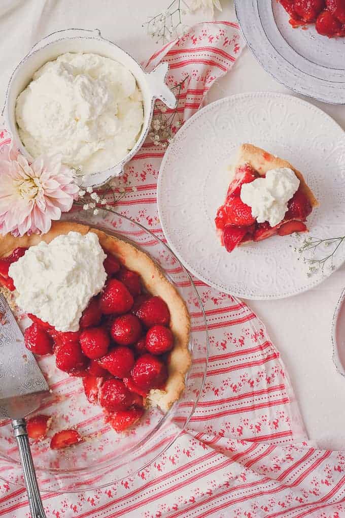 photo of fresh strawberry pie slice next to rest of pie, topped with whipped cream. there is a pink dahlia flower and a red and white table cloth.