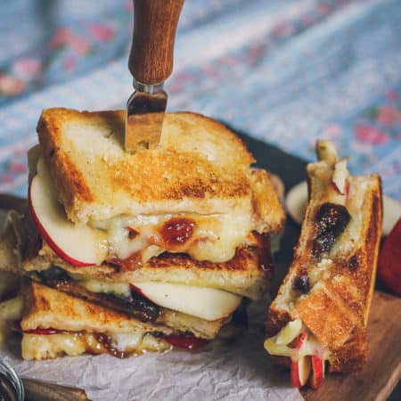 Image of stack of brie grilled cheese sandwiches with fig jam and apples. There's a cheese knife stuck into the top sandwich. There's another sandwich sitting upright next to the stack.